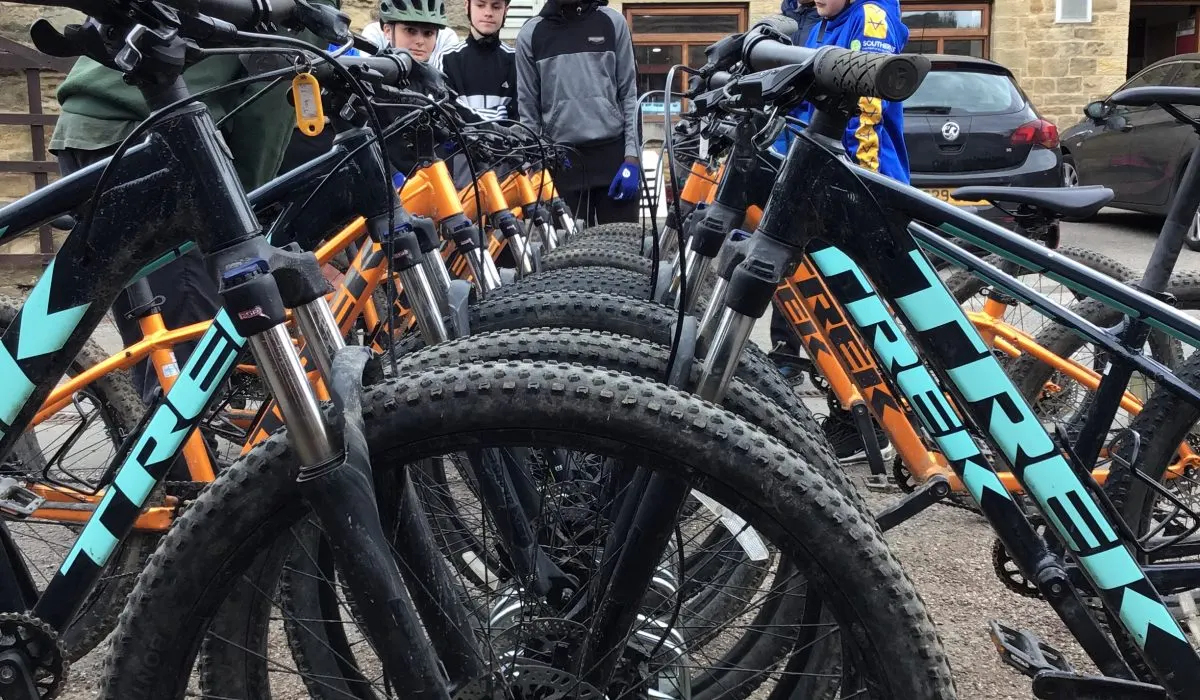 Row of orange and turquoise mountain bikes with fat tyres lined up in a courtyard, with visitors standing behind them.
