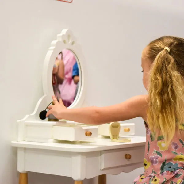 A young child with blonde hair touches an interactive mirror display on a white toy vanity unit.