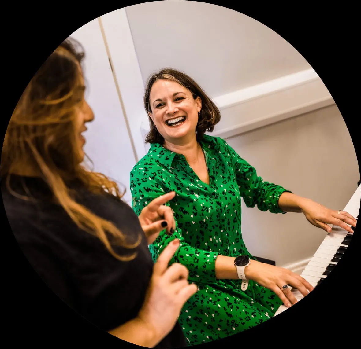 Two women enjoying a piano lesson together, one in a green patterned jacket smiling at the keys.