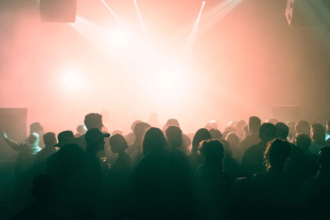 Crowd of people dancing and socializing in a nightclub with pink and green atmospheric lighting.