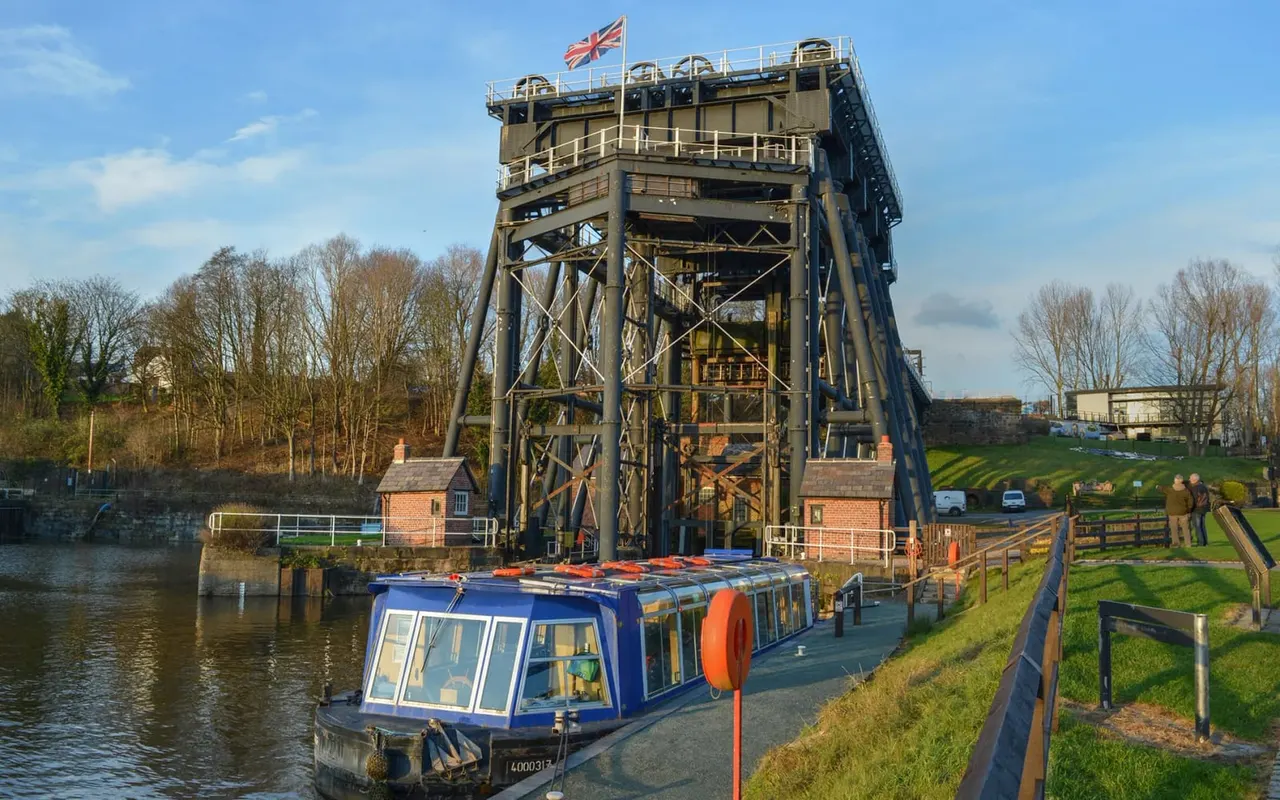 A blue and orange narrowboat moored beside a historic iron boat lift structure on a canal, with bare trees and green fields visible.