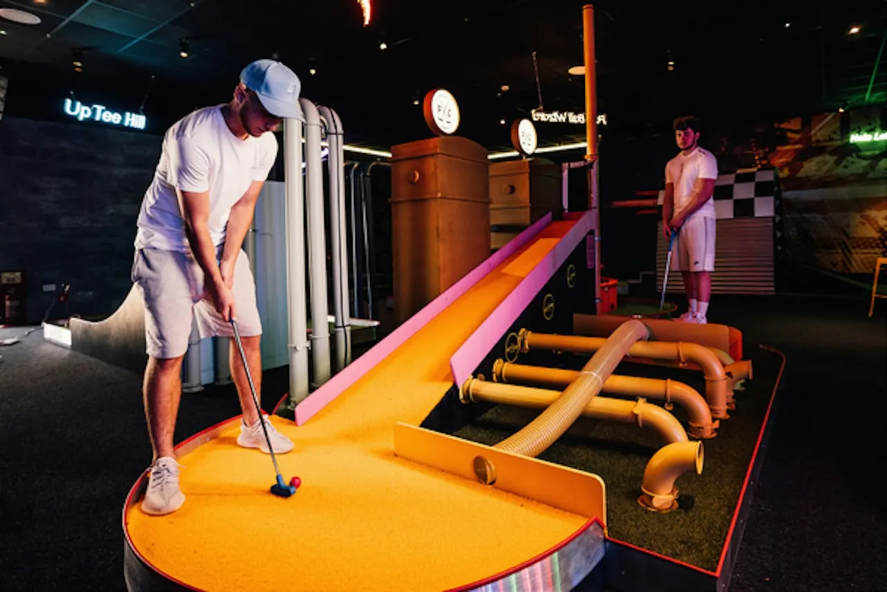 Two players enjoy an indoor crazy golf course with vibrant neon-lit obstacles and bright yellow and pink turf.