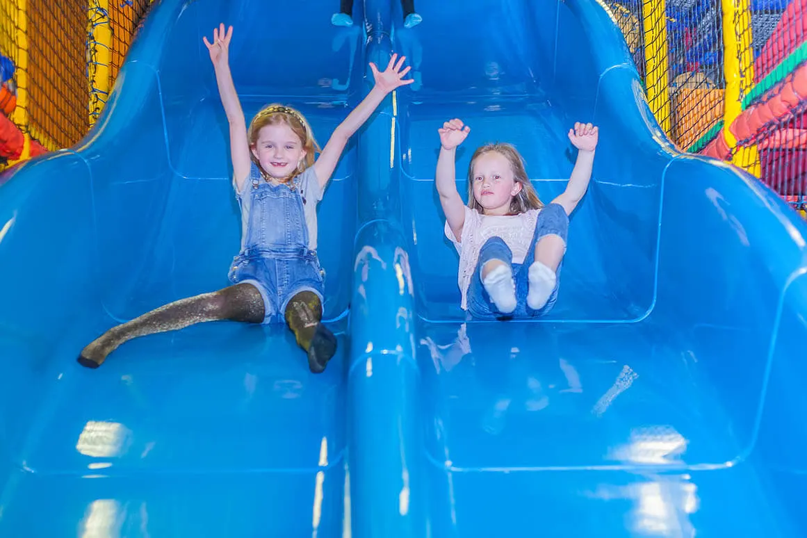 Two young children sliding down a large blue slide with arms raised, smiling at an indoor play centre.