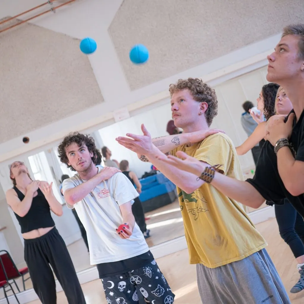 A group of young people juggling blue balls indoors during a circus skills workshop.