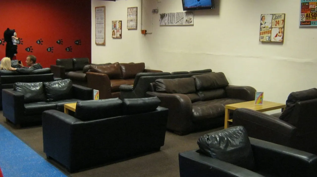 Spacious lounge area with multiple black leather sofas, wooden tables, a TV screen and vintage wall signs.