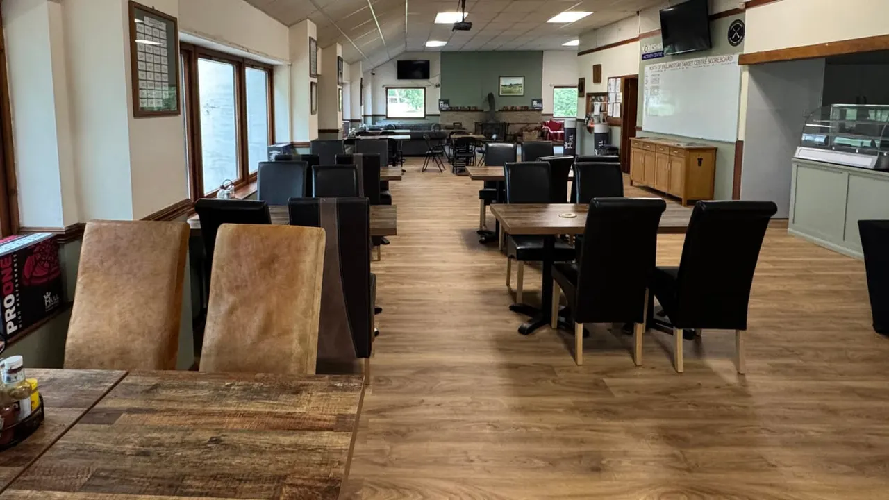 Spacious indoor hall with wooden flooring, dining tables with black and tan chairs, and activity equipment visible in background.