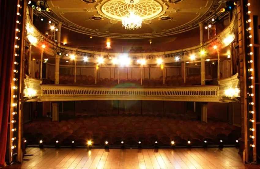 Empty historic theatre auditorium with ornate ceiling, grand chandelier, and illuminated balconies overlooking wooden stage.