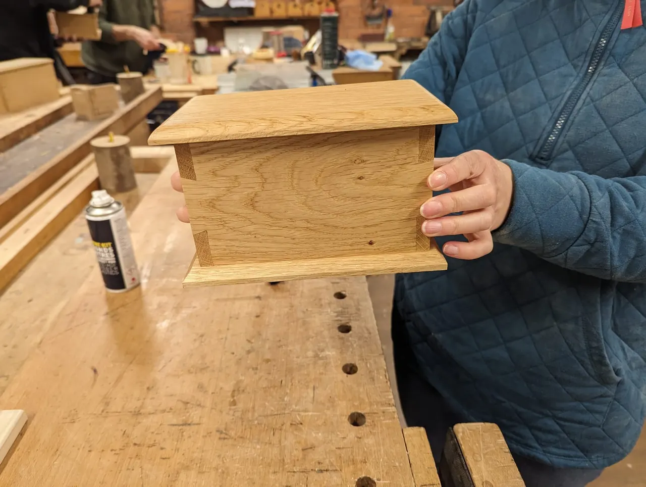 Person holding a finished wooden birdbox in a workshop with other participants and woodworking equipment visible in the background.