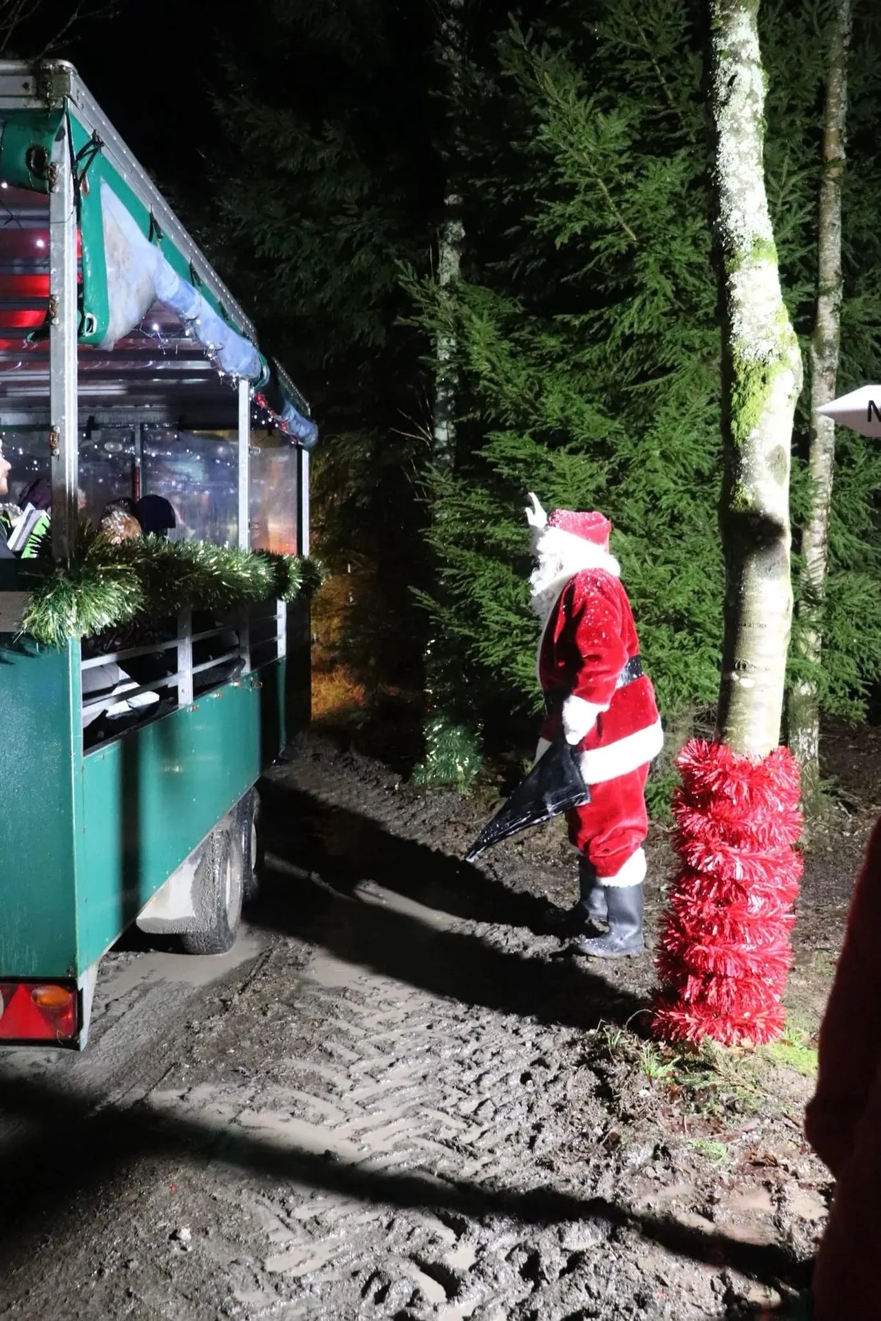 A figure dressed as Father Christmas stands at a festive market stall in woodland at night, surrounded by evergreen trees and red tinsel decorations.