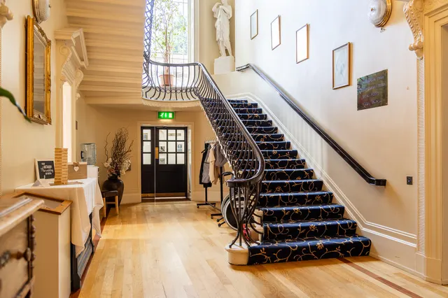Grand entrance hallway with wooden flooring, dark blue carpeted staircase, classical sculptures, and framed artworks on cream walls.