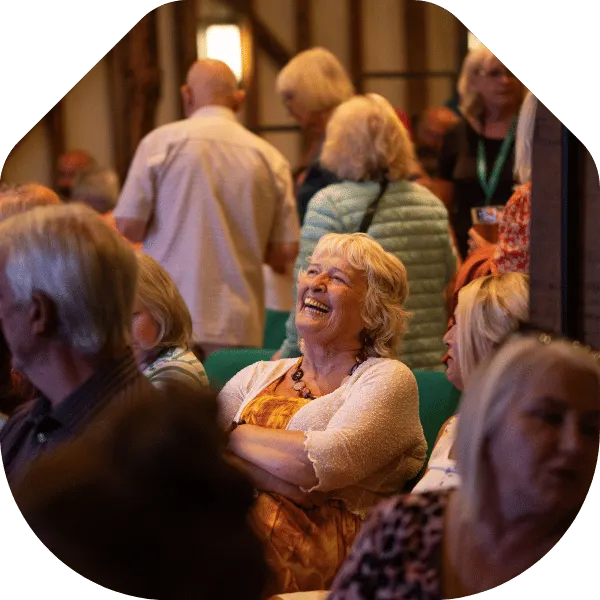 A smiling older woman laughs whilst socialising with other guests at an indoor event.
