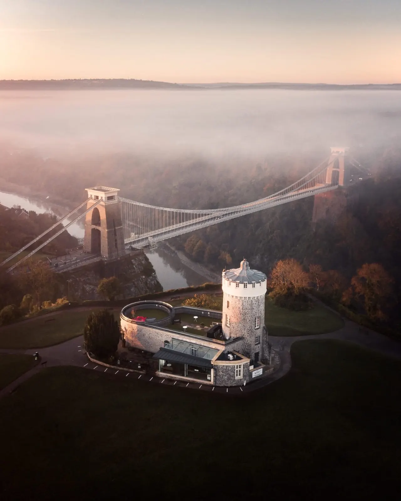 Aerial view of Clifton Observatory's round tower building with suspension bridge spanning misty gorge beyond.
