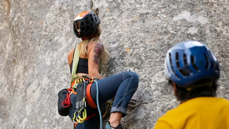 Person in orange and black helmet rock climbing an outdoor stone wall whilst wearing safety equipment, with another climber in yellow shirt watching nearby.