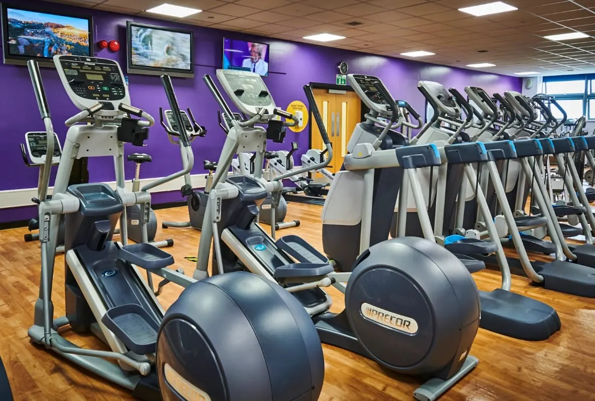 Gym floor with multiple exercise bikes and treadmills arranged in rows against purple walls with screens.