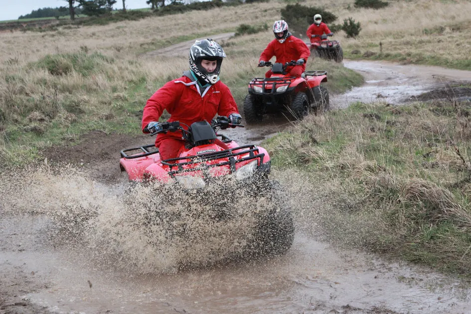 Three people in red jackets riding quad bikes through muddy terrain and water splashes.