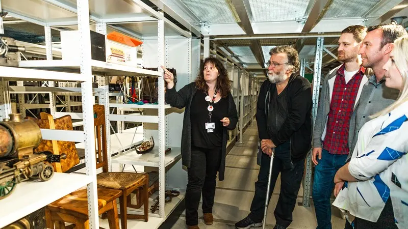 A woman in black leading a group of visitors through a museum storage area with wooden furniture and artefacts on shelves.