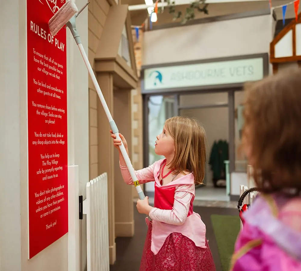 Young child in pink apron pointing at a red "Rules of Play" sign on an indoor play venue wall.