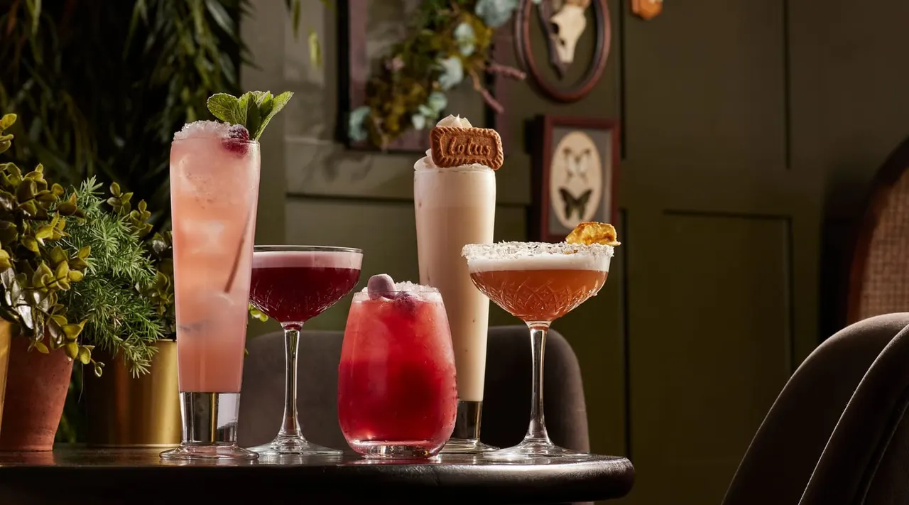 Five colourful cocktails displayed on a bar counter, garnished with mint, biscuits and other decorative toppings, with potted plants and artwork visible in the background.