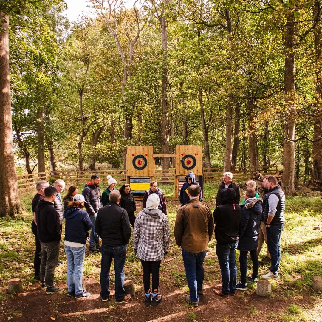A group of visitors standing in a woodland clearing around axe-throwing targets during an outdoor activity session.