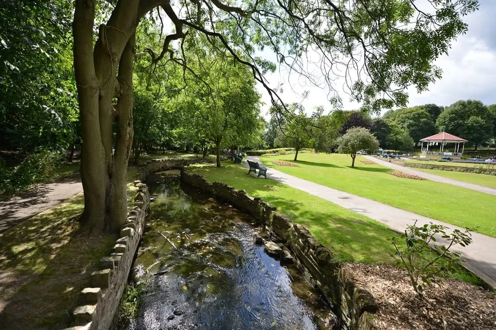 A stream flows through parkland bordered by mature trees, with open grass, a pathway and a gazebo visible in the distance.