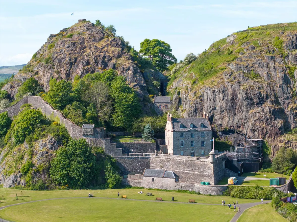 Stone castle nestled between dramatic rocky cliffs with visitors on the grassy grounds below on a sunny day.