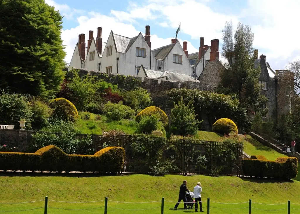 Two adults and a child in a pushchair walk across a lawn towards a historic white and stone manor house with distinctive chimneys, surrounded by manicured topiary hedges and castle ruins.