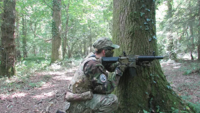 Person in camouflage and cap aiming an airsoft rifle whilst crouched behind a large tree in a wooded area.