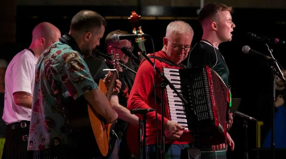 Four male musicians performing on stage with guitar, accordion and microphones under pink stage lighting.