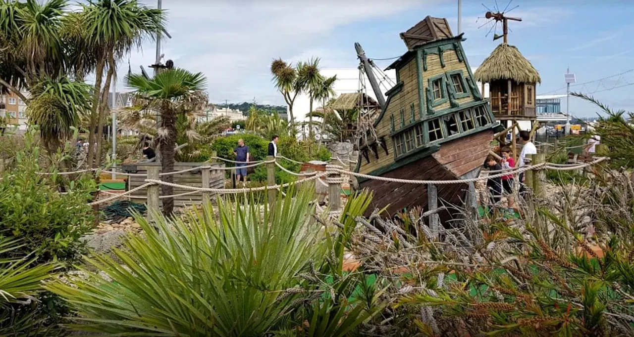 Visitors playing miniature golf on a themed pirate course with a tilted shipwreck building and tropical plants.
