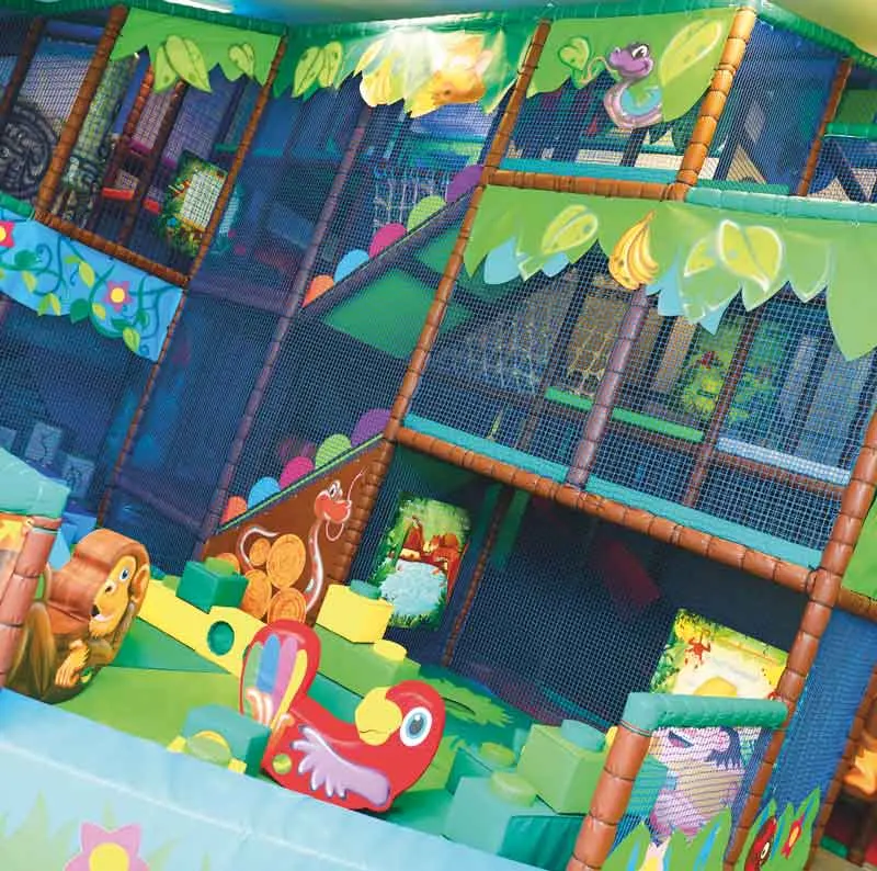 Overhead view of a colourful indoor soft play area with padded climbing structures, netting, and foam play equipment decorated with tropical fruit and animal designs.