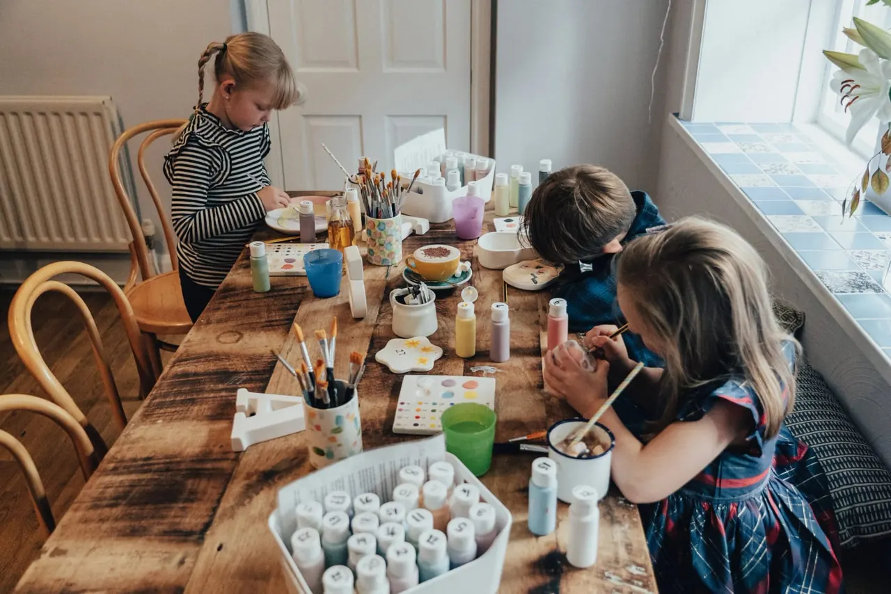 Children engaged in an art and craft activity at a wooden table with paints, brushes, and materials.