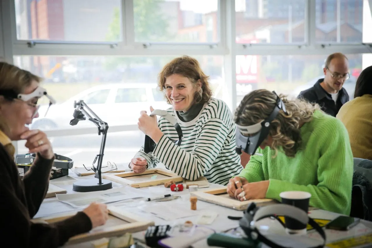A group of adults participate in a hands-on craft workshop at a bright, windowed studio table with tools and materials spread before them.