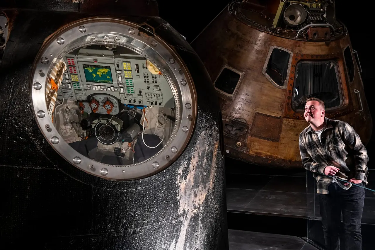 Man stands beside a vintage spacecraft capsule with circular hatch opening to reveal control panels and instruments inside.