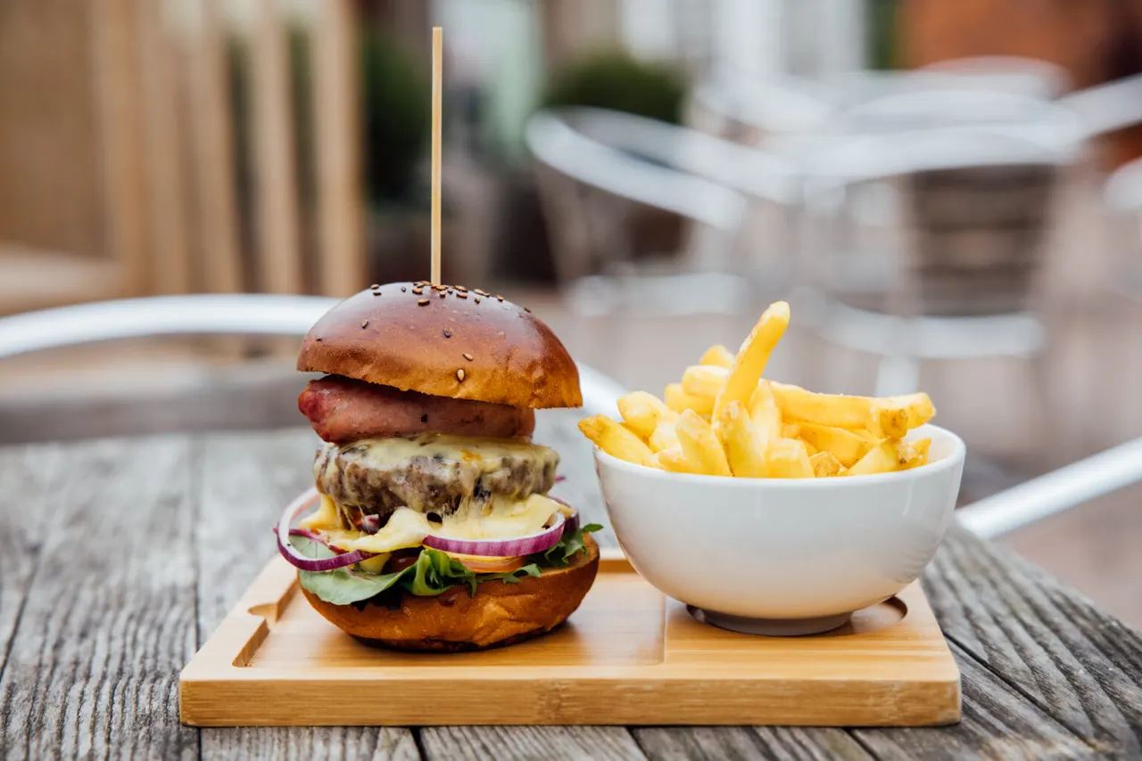 Gourmet burger with cheese and red onion served with chips in a white bowl on a wooden board at an outdoor terrace.