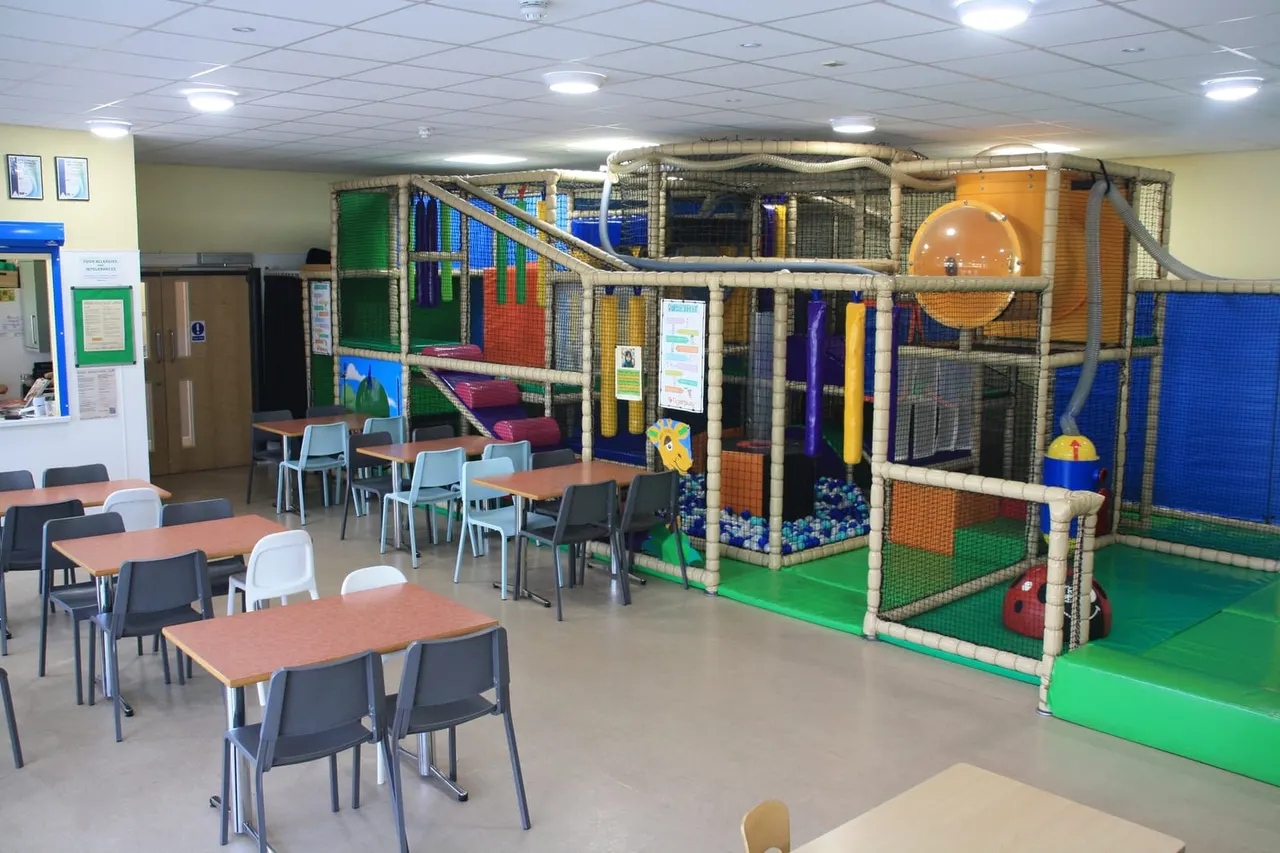 Indoor soft play area with dining tables, seating, and multi-level play structures with ball pits and slides.
