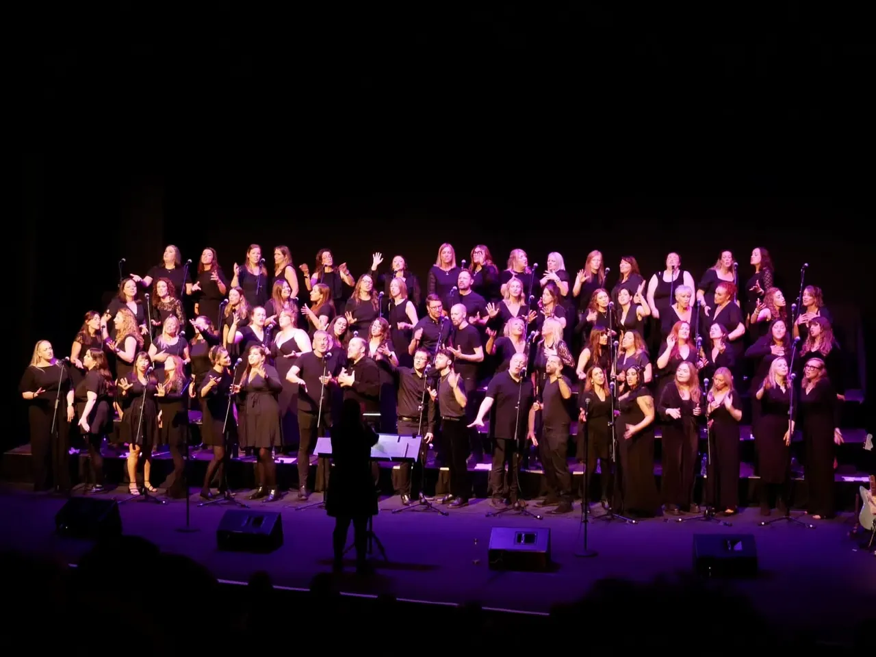 Large choir of around 60 singers in black clothing performing on stage under purple lighting with a conductor.