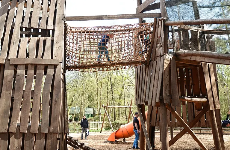 Child climbing through a rope net obstacle course at an outdoor adventure playground surrounded by wooden structures and trees.