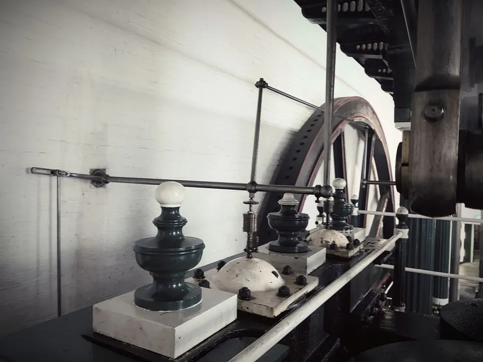 Historic beam engine machinery with cast-iron components and large wheel displayed indoors against a white brick wall.