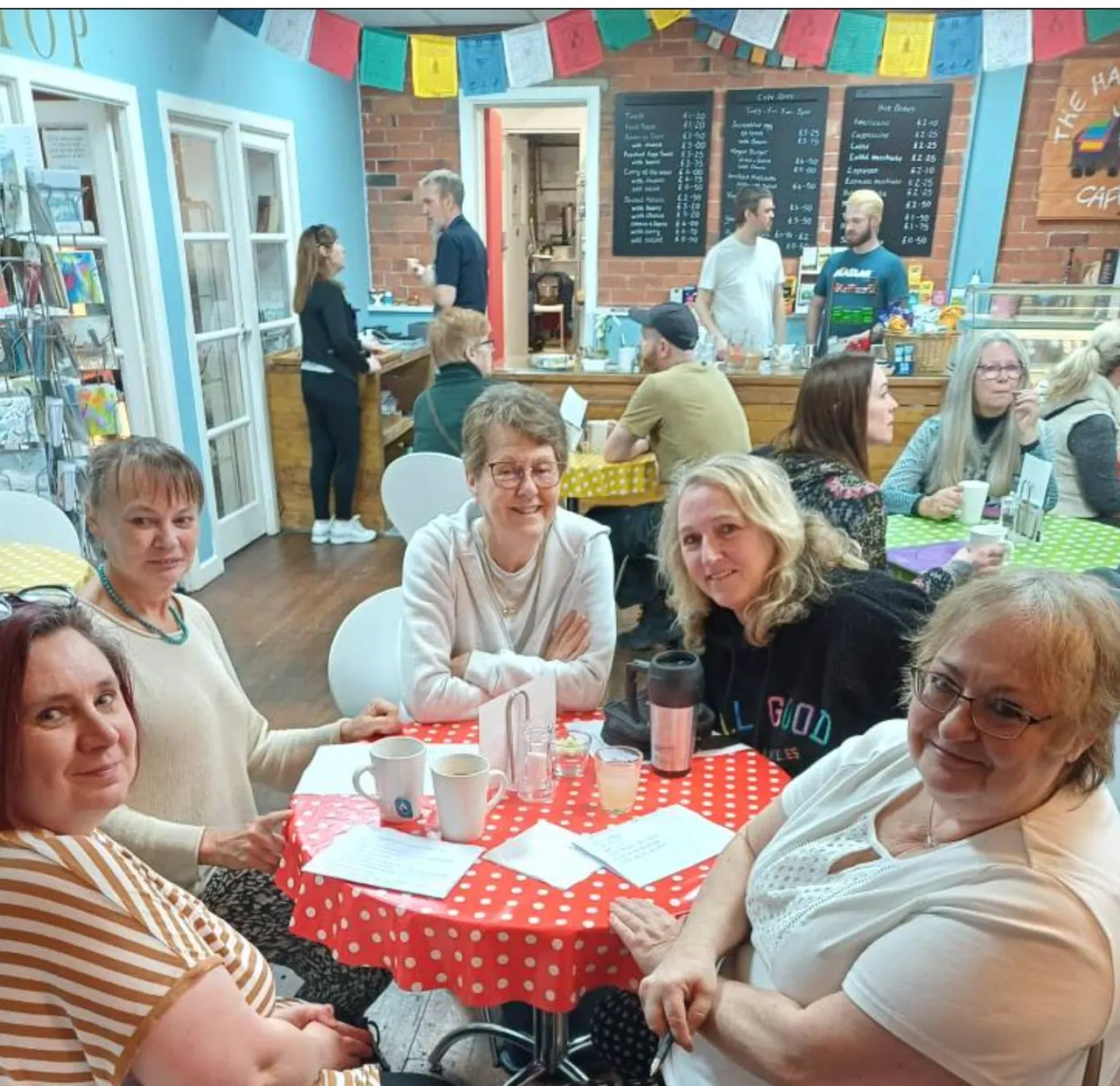 Groups of visitors enjoying refreshments and socialising at tables inside a vibrant community café space with colourful bunting and menu boards.