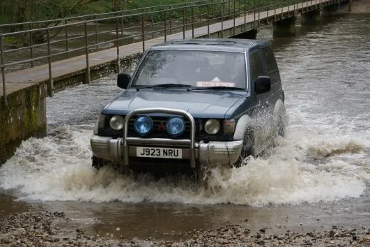 A four-by-four vehicle drives through deep floodwater, creating large splashes, beside a metal-railed footbridge.