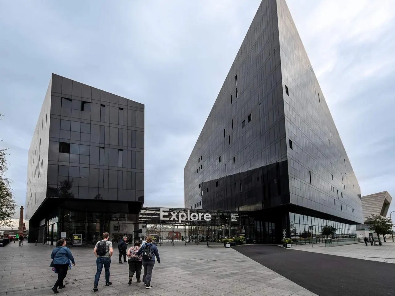 Visitors walk across a paved plaza in front of the modern Explore building with angular grey façade and large windows.