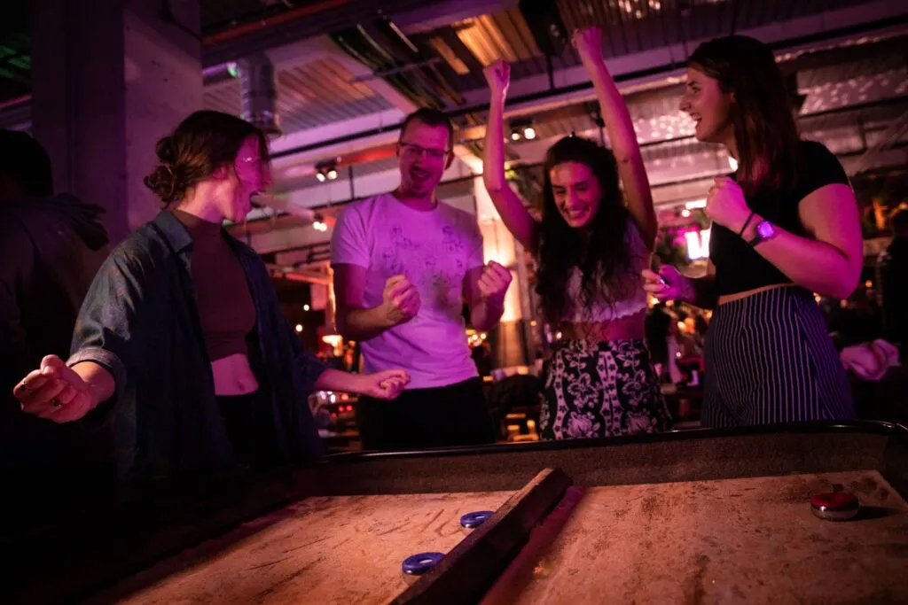 Group of five people playing air hockey and mini golf under vibrant purple and pink neon lighting in an indoor leisure venue.