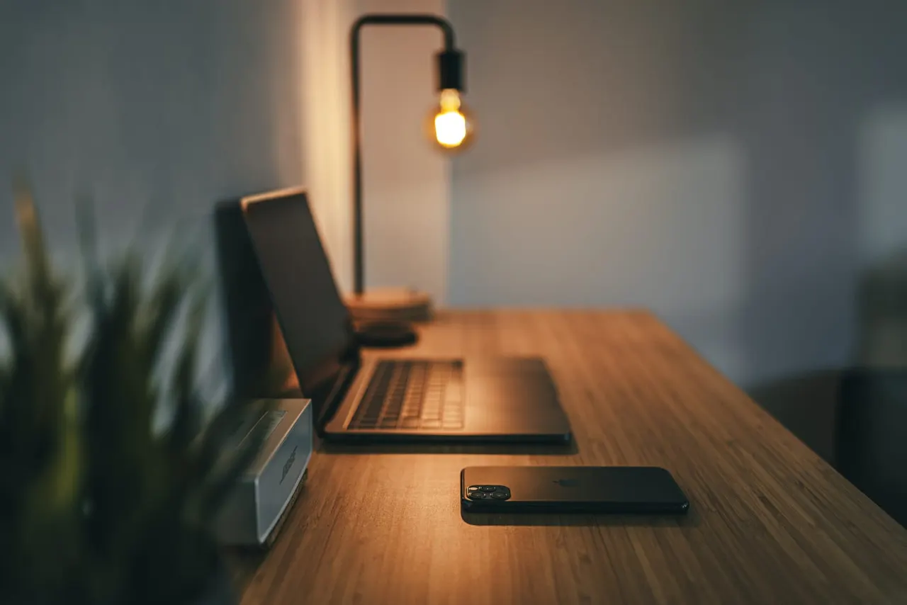 A wooden desk with a smartphone, laptop, and potted plant, lit by a warm pendant bulb.