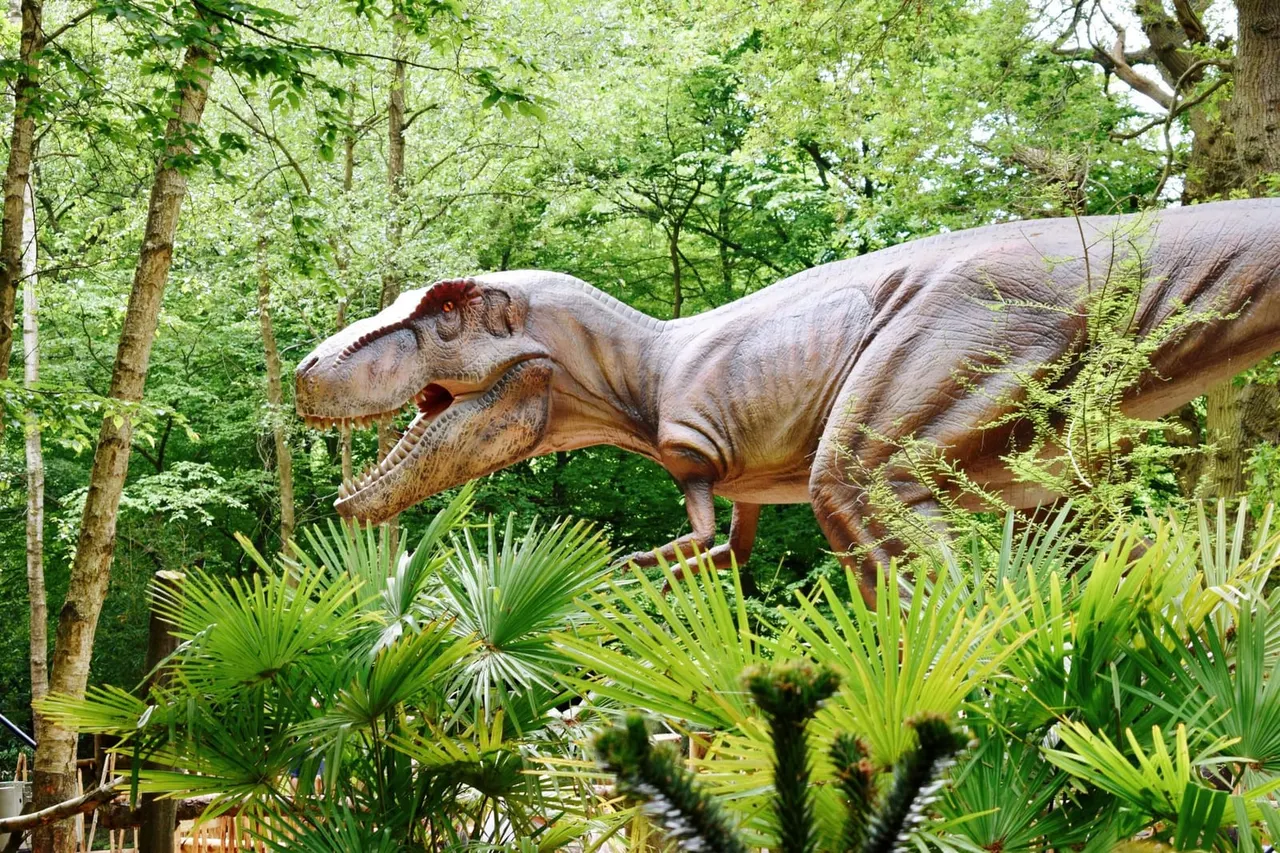 Life-sized dinosaur sculpture positioned among trees and tropical plants in a woodland setting.