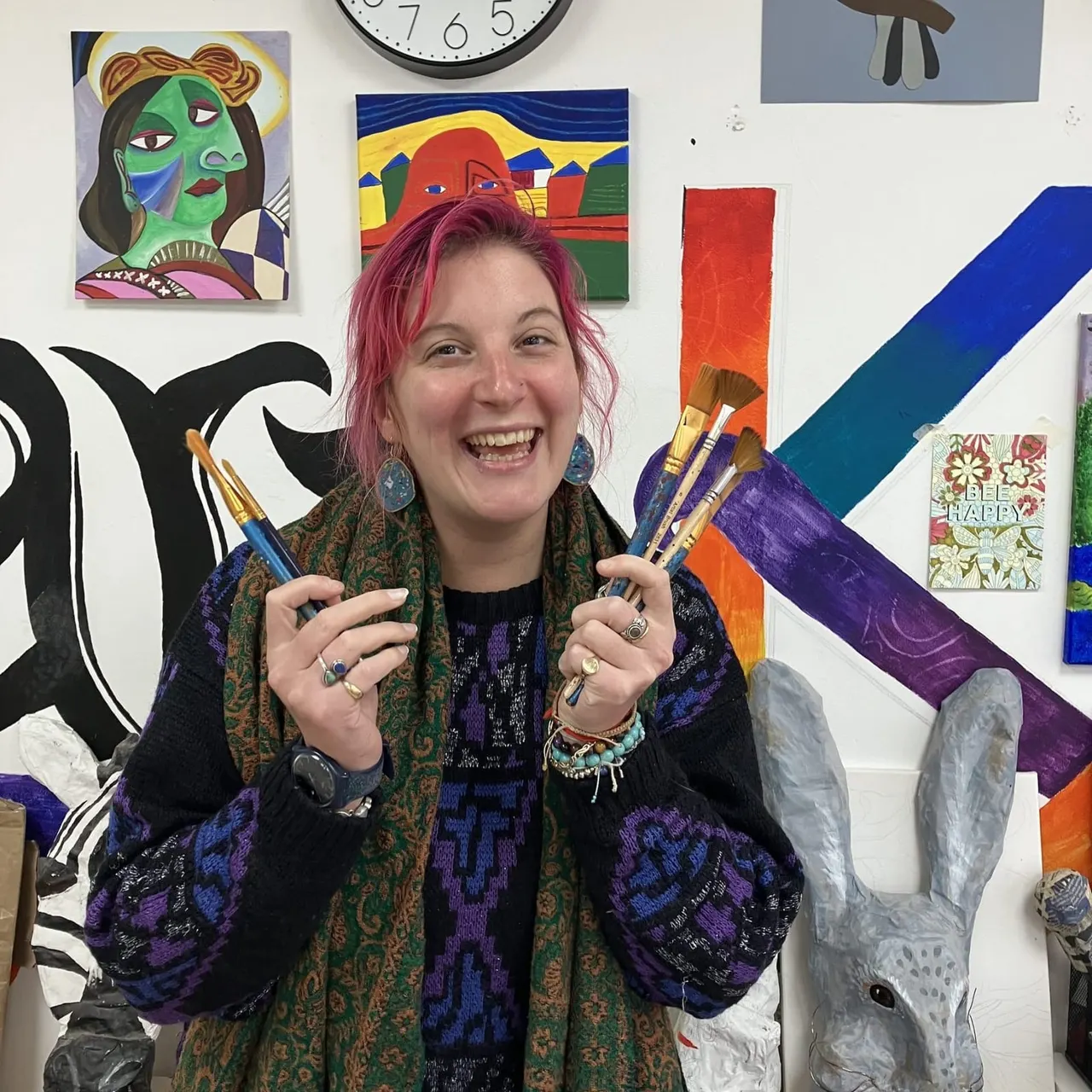 Woman with pink hair holding paintbrushes in an art studio surrounded by colourful artwork and creative decorations.