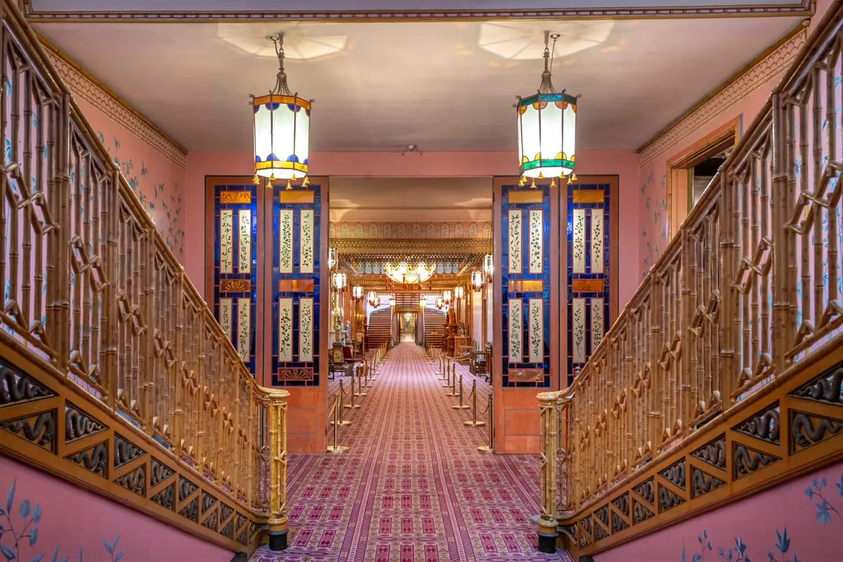 Symmetrical corridor with ornate wooden railings, pink walls, decorative blue and gold doors, and hanging lanterns in a grand historic interior.