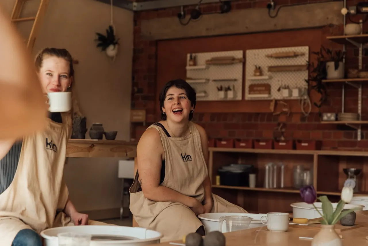 Two staff members in beige uniforms sit at a wooden table in a pottery studio, smiling amid ceramic pieces and shelving.