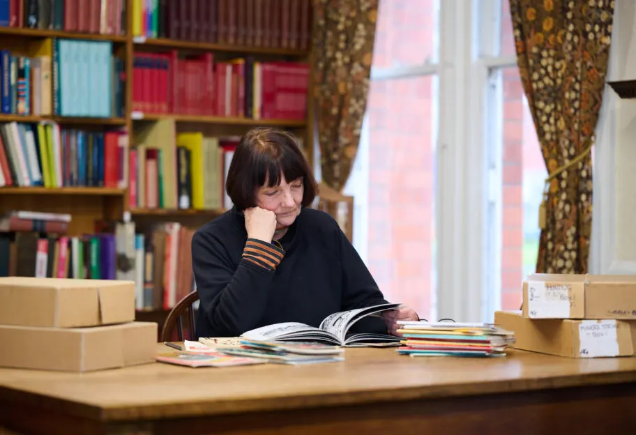 Woman reading a book at a wooden table surrounded by stacked books, with colourful bookshelves behind her.