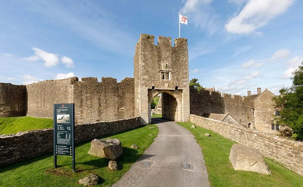 Pathway leading to the stone gatehouse of a medieval castle with crenellated walls and a flag flying above.