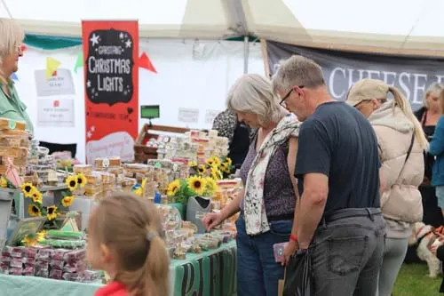 Visitors browse a market stall displaying produce and homemade goods under a canvas tent at an agricultural show.